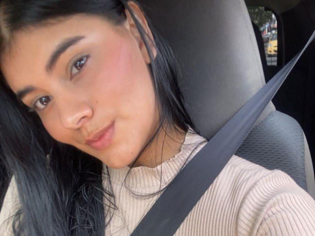 AlaraKey has black hair, brown eyes, and a slight smile. She's wearing a seatbelt, with a pink ribbed shirt, in a car, looking cute and relaxed.
