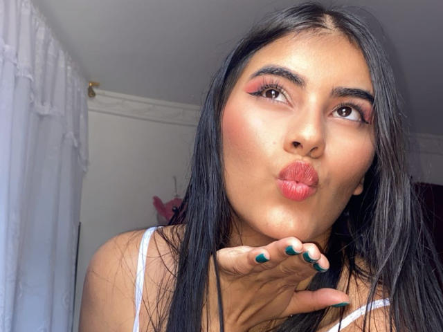 AlaraKey with long black hair, brown eyes, and pink lipstick, pouting her full, glossy red lips, touching her chin with green-painted nails. She's wearing a white top.