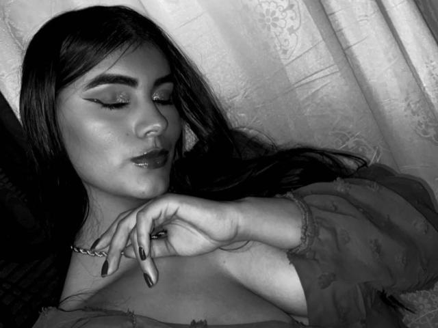 AlaraKey with dark hair, closed eyes, and glossy lips, wearing a sheer top, has her fingers near her face, showing long, manicured nails, and a chain necklace. Black and white.