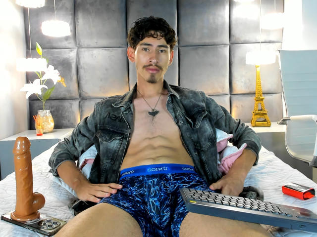 SkinnyJean, shirtless, wearing blue patterned underwear, sitting on bed with erect brown dildo, keyboard, and Eiffel Tower lamp in background, grey denim jacket, mustache, necklace, and relaxed expression.