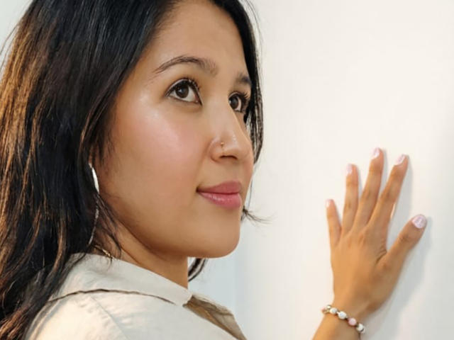 Het gezicht van LuxyLeim, met een medium bruine huid, donker steil haar, bruine ogen, roze lippenstift, een kleine neusring, een wit shirt, en een hand tegen een witte muur, waarbij roze nagellak te zien is..