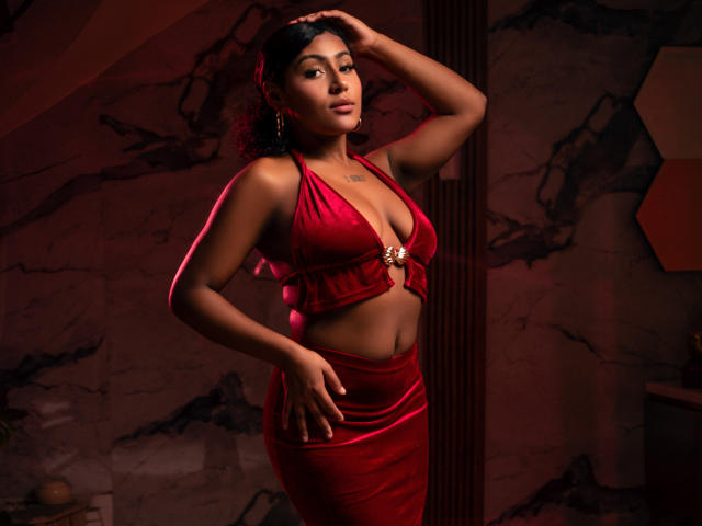 AmberrQueen met donker krullend haar, gekleed in een rode satijnen topje met halter en een bijpassende rok, waarbij haar borsten en billen zichtbaar zijn, met haar hand in haar haar, rood licht, zelfverzekerde pose, gouden oorbellen..
