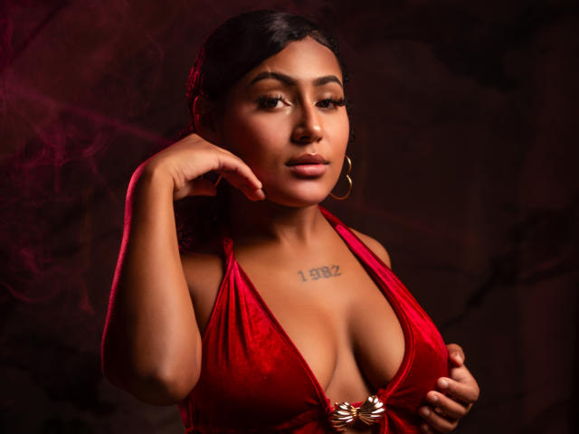 AmberrQueen, avec ses cheveux foncés, portant un haut en velours rouge à encolure dégagée, montrant ses seins volumineux, une main près de son visage, des boucles d'oreilles dorées, un tatouage au-dessus de sa poitrine, sexy, fond sombre..