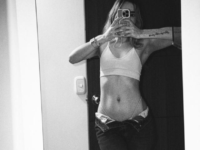 Autoportrait en noir et blanc d'AdharaCooperr avec des cheveux blonds, portant un soutien-gorge de sport, un jean déboutonné et des sous-vêtements visibles, tenant un téléphone, montrant un bras tatoué..