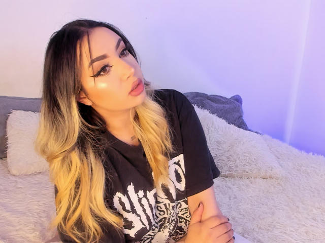 MelissaLust with blonde and black ombre hair, wearing a black "zombie" t-shirt, has pouty pink lips and eyeliner, sitting on a fluffy white bed, looking at camera.