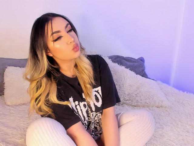 MelissaLust with blonde-to-black ombre hair, pouty lips, wearing a black t-shirt and white ribbed pants, sitting on a fluffy white bed, looking sexy.