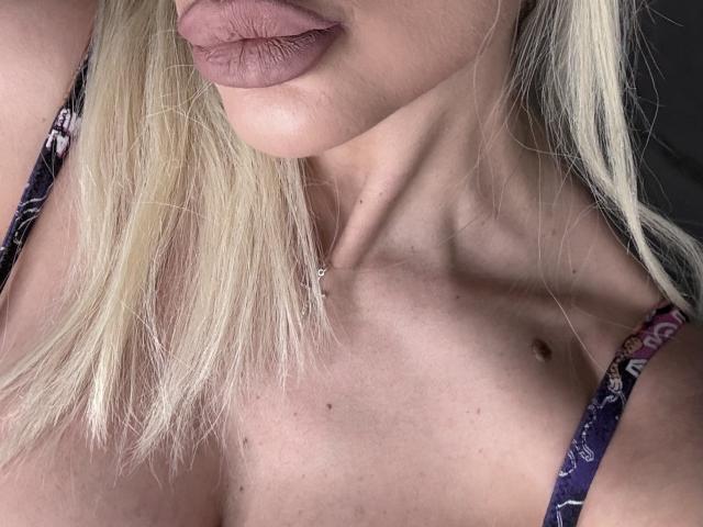 Les lèvres charnues et rosâtres de Lellie sont légèrement entrouvertes. Elle a les cheveux blonds, un teint clair et une tache de naissance visible sur la poitrine. Elle porte un haut violet à motifs..