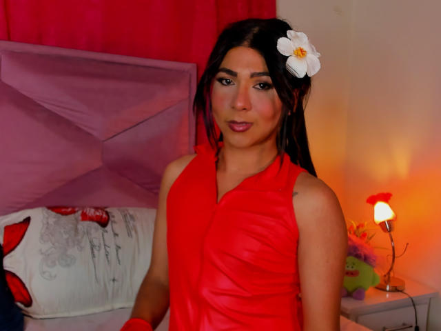 ZoeHotMoretti, com cabelos longos e escuros, vestindo um vestido de couro vermelho com alças, com uma flor no cabelo, olhando para a câmera, com um sorriso leve, batom vermelho, cama com travesseiros rosa e brancos ao fundo..