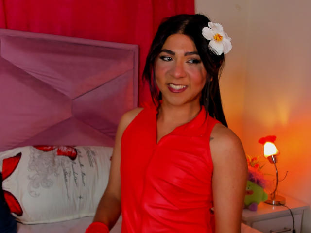 ZoeHotMoretti met lang zwart haar, rode mouwloze jurk, witte bloem in het haar, lachend, rode make-up, zittend op een bed, lichte huid, kleine borsten, schattig, warme verlichting in de slaapkamer..