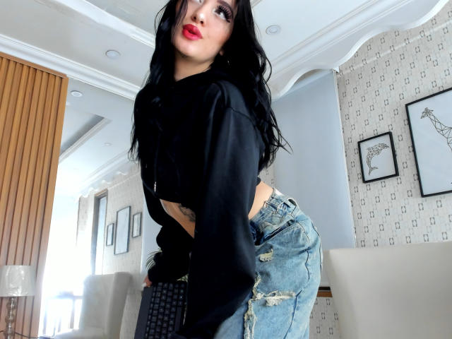 Tifany Diaz mit schwarzen Haaren, rotem Lippenstift, einer schwarzen Kapuzenjacke und zerrissenen blauen Jeans, die nach vorne gebeugt ist und ihr Gesäß zeigt, in einem modernen Raum mit Holzwänden und gemusterten Wänden..