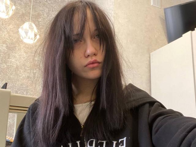SantinaSahm with messy black hair, medium tits, wearing a black hoodie, looking slightly bored, in a bright room with geometric hanging lights.