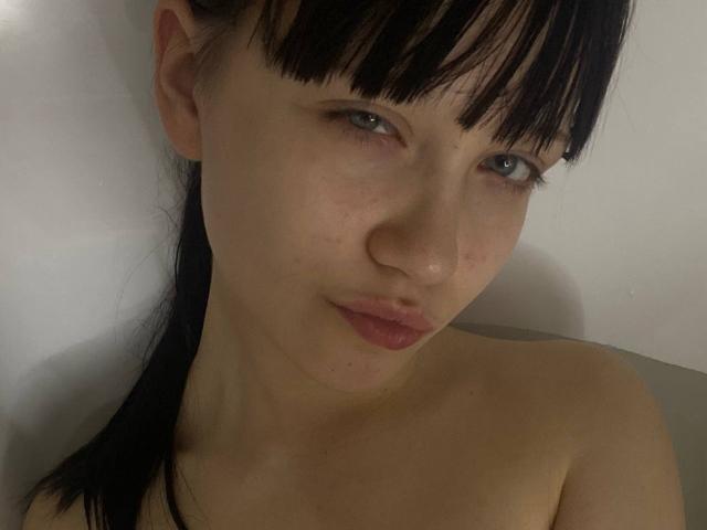 SantinaSahm has pale skin, black hair with bangs, blue eyes, and is topless, showing her small, bare boobs. She's looking at the camera with a slight pout.