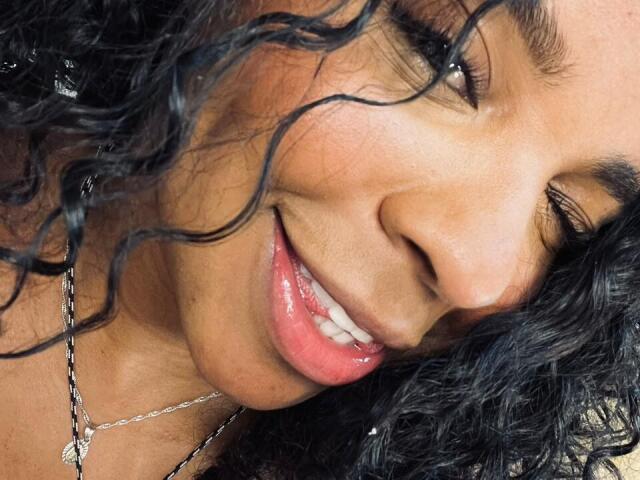 LisaMendez's face with curly black hair, brown skin, and a slight smile showing her pink tongue. She's wearing a silver necklace with a small pendant.