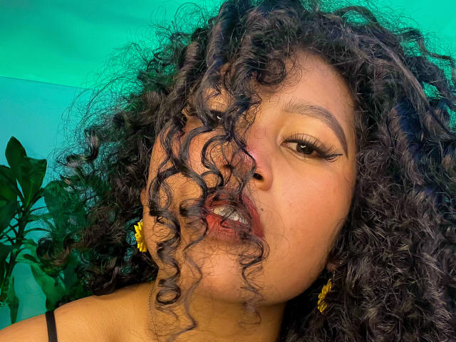 SophieSonyas con i capelli neri ricci, orecchini a forma di fiore giallo e eyeliner nero, leggermente con le labbra arricciate e leggermente aperte, mostrando i denti. Sfondo verde..