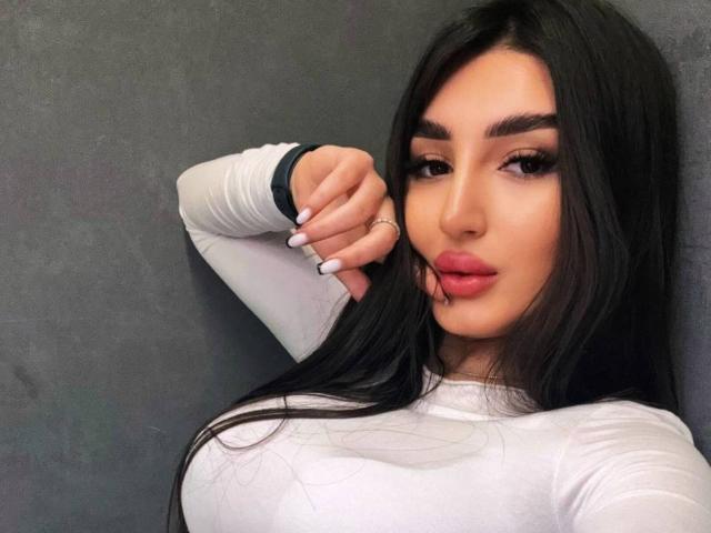 EllynBousqute with long black hair, full lips, and white long-sleeve top, showing off her tits, hand raised, dark eyebrows, and white nails.