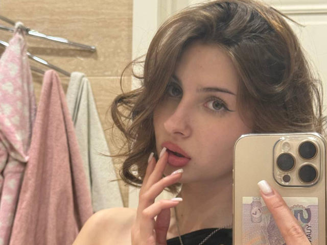 NellieHeimer with wavy brown hair, light skin, and winged eyeliner, sucking her finger, holding a gold iPhone, pink and gray towels in the background.