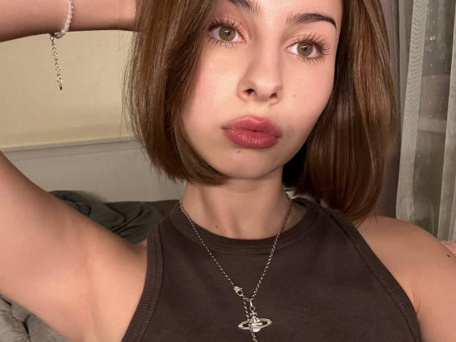 NellieHeimer with brown bob, green eyes, and pink lips, wearing a brown sleeveless top and silver necklace, arm raised, looking at camera, slightly pouty expression.