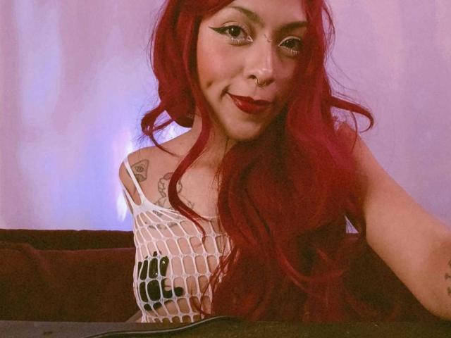 De aspecto sensual, con cabello rojo, vistiendo una camiseta blanca de rejilla que deja ver sus pechos, tiene tatuajes en el hombro y un piercing en la nariz, sonriendo con lápiz labial rojo en un selfie..