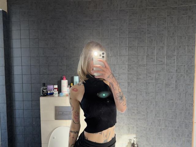 SensualMila with blonde hair, black sleeveless top, tattoos on arms and side, taking mirror selfie in bathroom, phone flash in face, gray tiled wall, toiletries on sink.