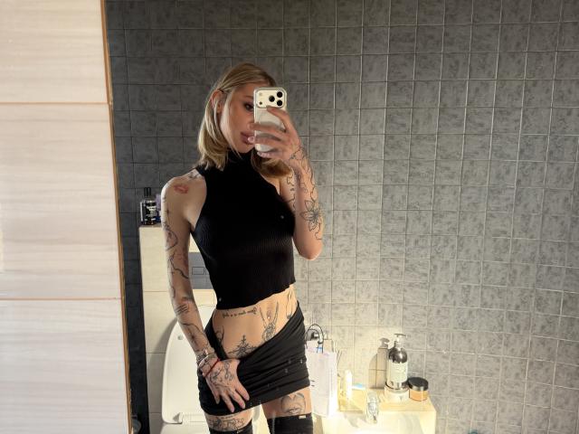 SensualMila, avec ses cheveux blonds, son haut noir sans manches, sa mini-jupe noire, ses tatouages sur les bras et le ventre, tenant un téléphone, dans un selfie devant le miroir de la salle de bain, montrant ses fesses et ses tatouages..