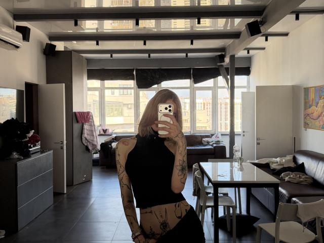 SensualMila, avec des tatouages, un débardeur noir et une jupe noire, debout dans une pièce, tenant un téléphone, se cachant les seins, montrant ses fesses, et un tatouage visible sur son bras..