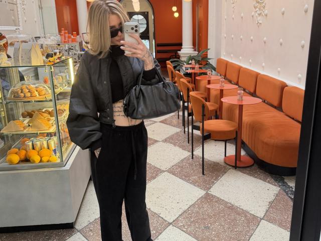 SensualMila avec une coupe de cheveux blonde, des lunettes de soleil noires, une veste en cuir, un pantalon noir et un ventre légèrement visible, tenant un téléphone, un sac noir, debout dans une boutique de beignets, prenant un selfie devant un miroir..