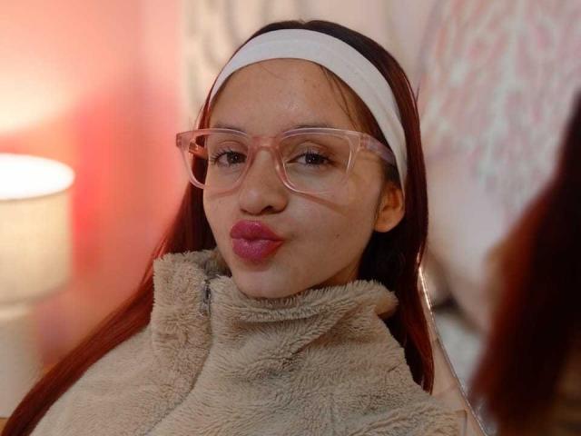 LesleyGray, avec des lunettes roses, des lèvres roses et pulpeuses, des cheveux bruns, un bandeau blanc, un pull en cachemire beige et un éclairage rouge doux, avec une lampe blanche en arrière-plan..