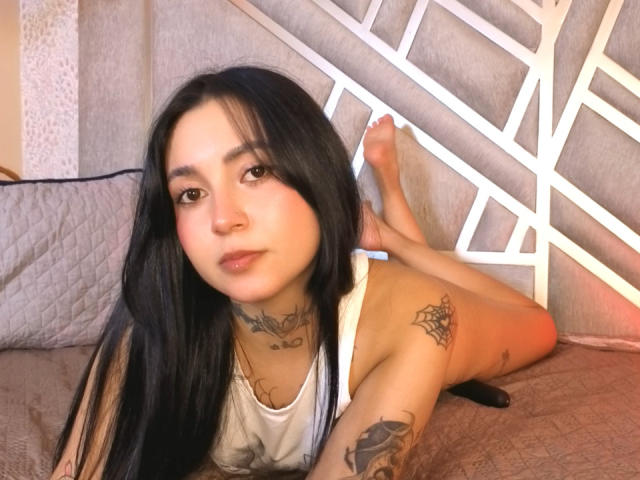 GabyMathew with long black hair, lying on a bed, wearing a white tank top, showing tattoos on neck, chest, and arm, legs up, bare feet, looking at camera.