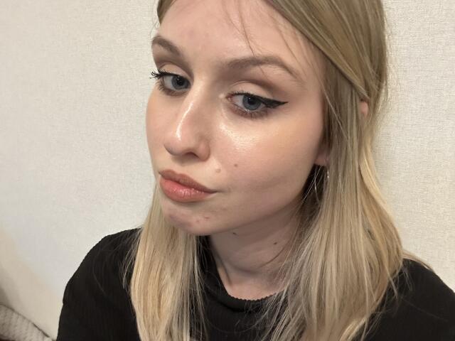 Nikollia ha una pelle luminosa, capelli biondi, eyeliner nero e un'espressione neutra. Indossa una maglia nera con una leggera texture e guarda in basso, con labbra di un delicato rosa..