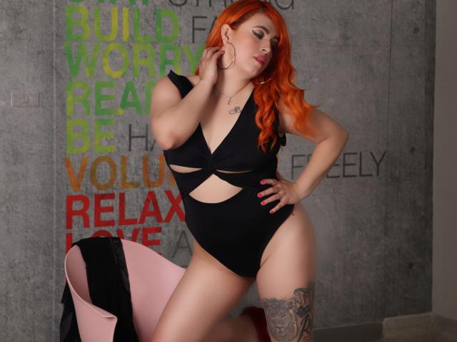 Lilibel con i capelli rossi, indossa un costume intero nero con tagli che mostrano il seno e i glutei, ha un tatuaggio sulla coscia, è seduta su una sedia rosa, c'è un muro grigio, ha una mano appoggiata al fianco, guarda in basso, ha le unghie rosa..