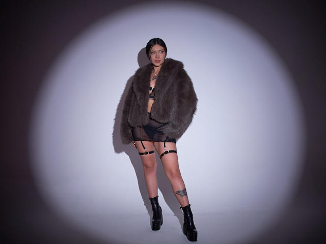 BerlyVenuss wears a gray fur jacket, black lingerie, garter, and high heels. She has tattoos on her thigh and leg, and a black choker. Her ass is visible.