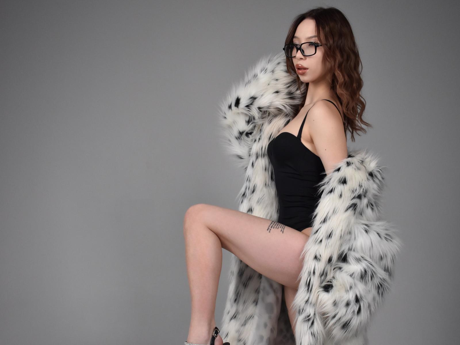 AshleyeGrey with glasses, black tank top, white and black furry coat, one leg up, tattoo on thigh, brown wavy hair, sexy, pale skin, slim, small tits, cute.