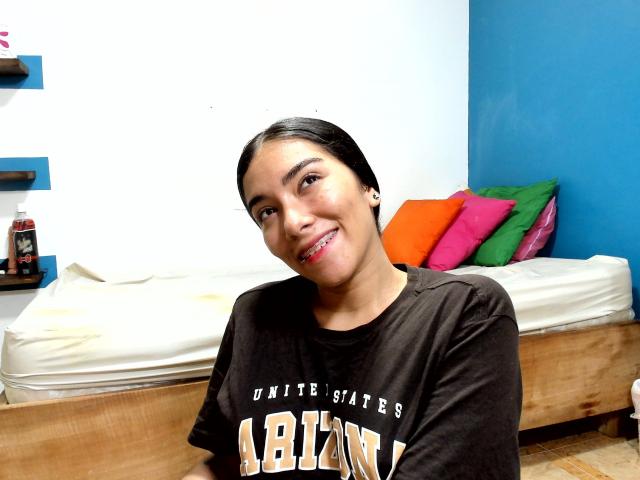 SharonKlart with black hair, smiling, wearing a brown "ABLIN" shirt, has small tits, and a cute face, standing in front of a bed with colorful pillows.