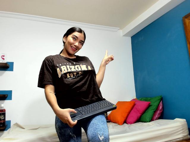 SharonKlart, smiling, black t-shirt, "Bizon 579-P", blue jeans, holding laptop, right index finger raised, white and blue walls, colorful pillows, black hair, small tattoo on left forearm.