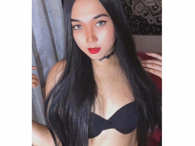 KateMadness met lang zwart haar, rode lippenstift, een zwarte bh en een choker, die naar de camera kijkt en haar borsten laat zien, in een slaapkamer met grijze gordijnen en een rood bed..