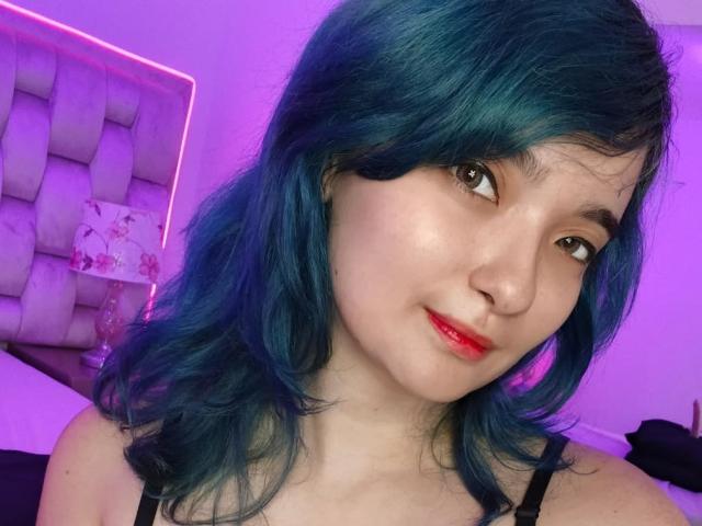 Naommy Carter, com cabelo azul, batom vermelho e um sorriso adorável, exibindo seus seios em uma regata preta, sob iluminação roxa..