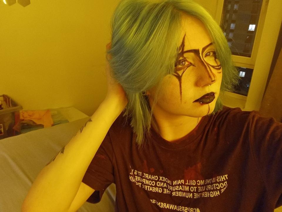 LouellaDupaski with green hair, black lipstick, and eye makeup, wearing a brown shirt, looking to the side, hand in hair, yellow lighting.