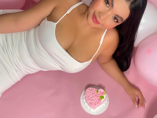 AlisBella with large tits, wearing a white dress, lying on pink, smiling at the camera, next to a pink heart-shaped cake with a rose and green leaf.