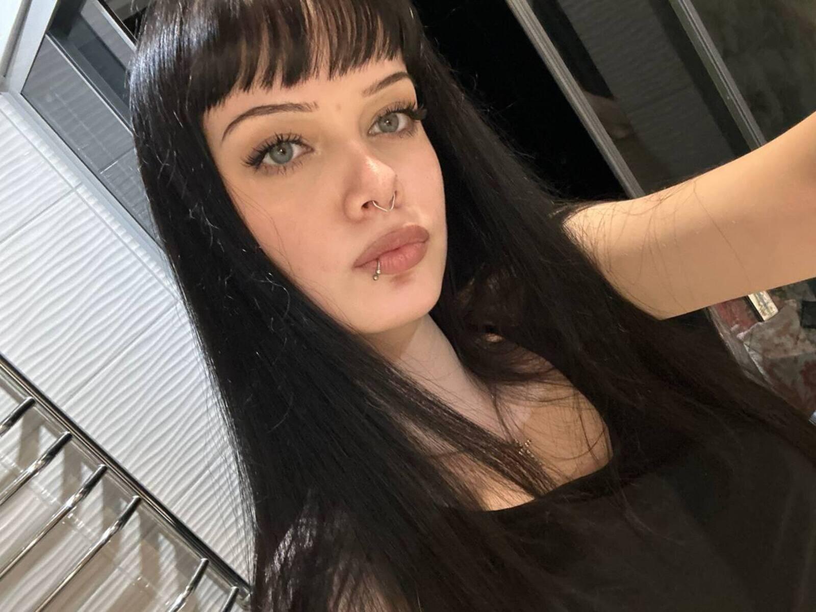 LizethTilbury with long black hair, pale skin, green eyes, nose ring, and lip piercing, wearing a black top, looking at the camera with a slightly pouty expression.