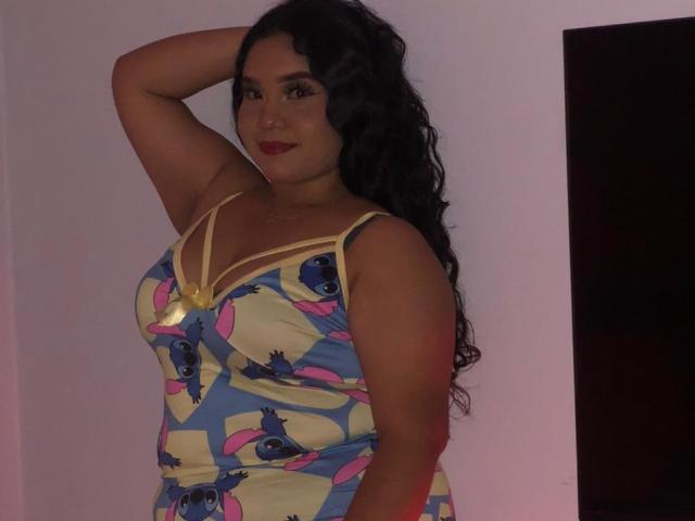 EvaSofia, con su largo cabello negro y rizado, vestida con un vestido colorido con patrones de ojos en azul, rosa y amarillo, y tirantes amarillos, tiene un brazo levantado, mostrando sus senos..