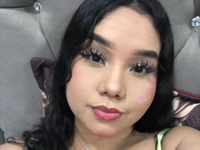 EvaSofia tem cabelos pretos longos, cílios grossos, batom rosa, um piercing no nariz, uma pequena espinha rosa na bochecha e está olhando para a câmera com um leve sorriso..
