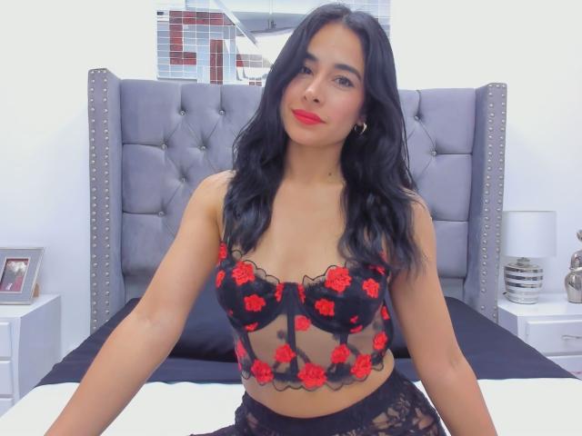 LyraRaven with long black hair, red lipstick, wearing a black lace and red rose lingerie top, showing her tits, sitting on a bed with a gray tufted headboard.