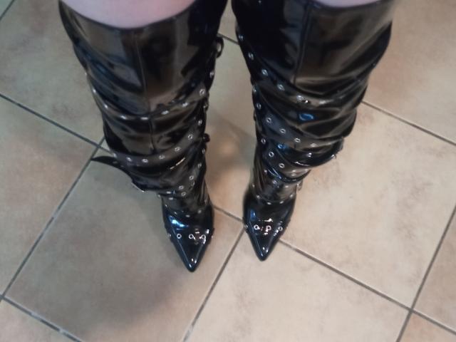 MonicaMiss's shiny black thigh-high boots with multiple buckles and pointed toes, standing on tiled floor. Boots are glossy and reflective, showcasing her plump, pale thighs.
