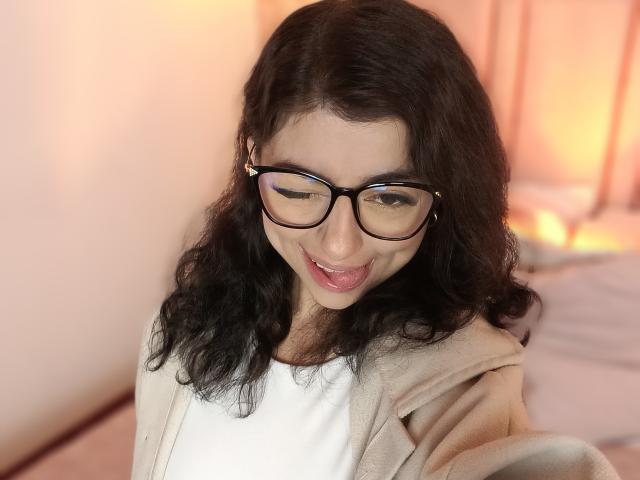 Anntonella Watson, met krullend zwart haar, zwarte bril en een schattige glimlach, die haar tong uitsteekt, draagt een beige vest over een witte top, in een zacht verlichte kamer..
