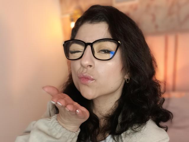 Anntonella Watson mit Brille, schwarzen, welligen Haaren, vollen, rosafarbenen Lippen und einer erhobenen Hand, die einen Kuss macht, trägt einen beigen Pullover und hat einen hellen Hautton..
