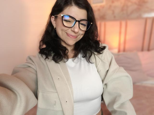 Anntonella Watson, avec ses cheveux noirs ondulés, ses lunettes et un cardigan beige clair par-dessus un haut blanc, sourit doucement, les bras tendus, dans une pièce confortable avec un éclairage chaleureux..