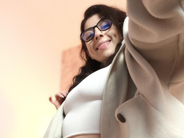 Anntonella Watson mit Brille, lächelnd in die Kamera, trägt einen beigen Mantel über einem weißen Sport-BH, wobei ihre Brüste sichtbar sind. Aufnahme aus niedriger Perspektive, mit Fokus auf ihr Gesicht und ihren Mantel..