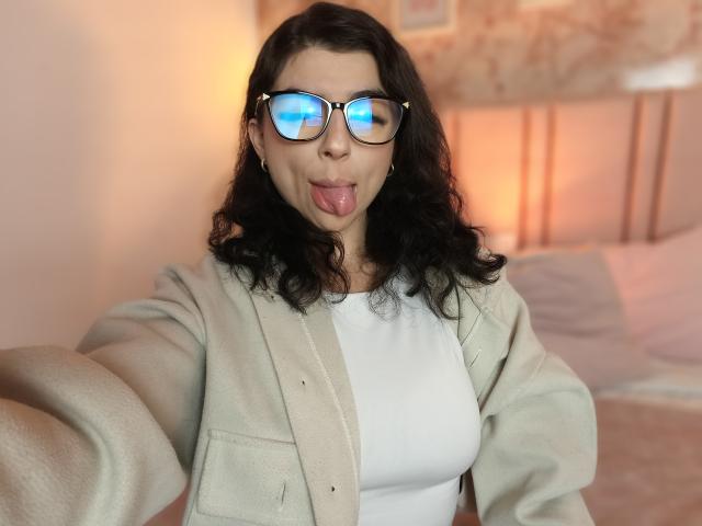 AnntonellaWatson with wavy black hair, wearing glasses reflecting blue light, sticking out her tongue, in a white top and beige cardigan, showing her tits.