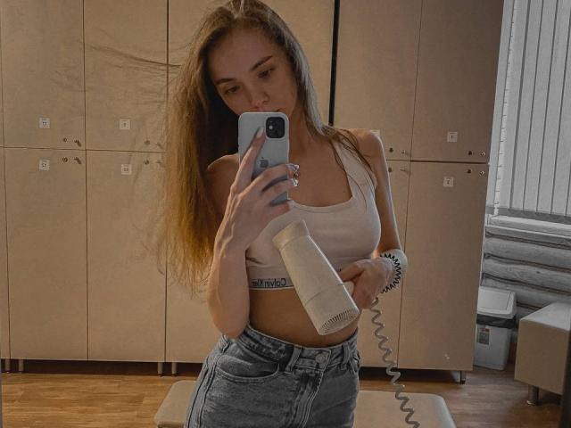 MirellaBagne mit langen, braunen Haaren, die einen Föhn in einem Spiegel-Selfie hält, trägt ein weißes Tanktop, das ihre Brüste zeigt, und blaue Jeans, in einem Umkleideraum..