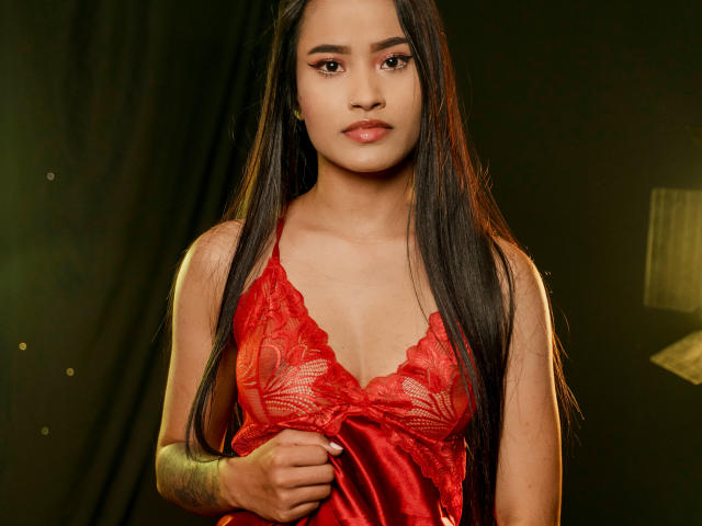 KarolPalacios with long black hair, wearing a red lace lingerie top, has small tits, and a tattoo on her left arm, standing against a dark green background.